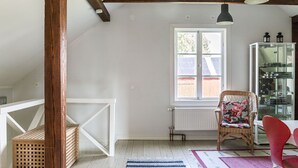 Ett rum med ett vitt tak, träbjälkar, ett stort fönster, en flätad stol, en röd stol, ett glasskåp och en blå- och vitrandig matta.
