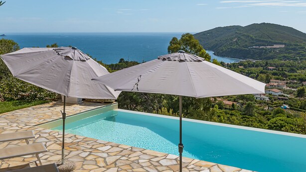A pool with stone tiles and lounge chairs, two umbrellas, and a view of the sea and hills. - Villa di Sogno by Interhome (Lacona)