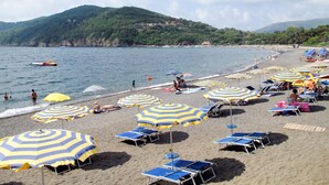 A beach with multiple sun umbrellas and lounge chairs, a sandy shore, and a view of the sea and hills in the background. - Villa di Sogno by Interhome (Lacona)