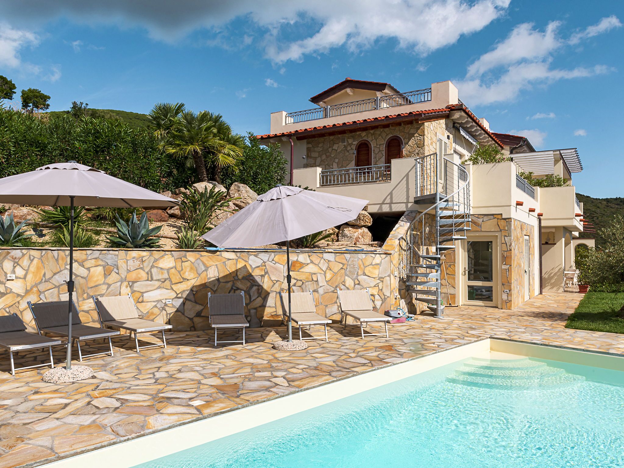 A pool area with stone flooring, lounge chairs, and umbrellas.