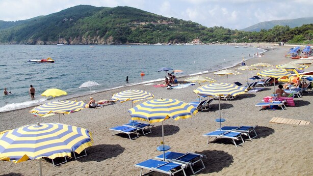 A beach with multiple sun umbrellas and lounge chairs, a sandy shore, and a view of the sea and hills in the background.