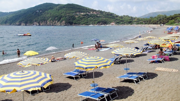 Une plage dotée de nombreux parasols et de chaises longues, une rive sablonneuse, et une vue sur la mer et les collines en arrière-plan.