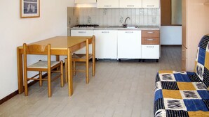 Une cuisine compacte avec des meubles blancs, une cuisinière à gaz et un évier. On y trouve une table à manger en bois avec des chaises, ainsi qu’un canapé à motifs dans l’espace salon.
