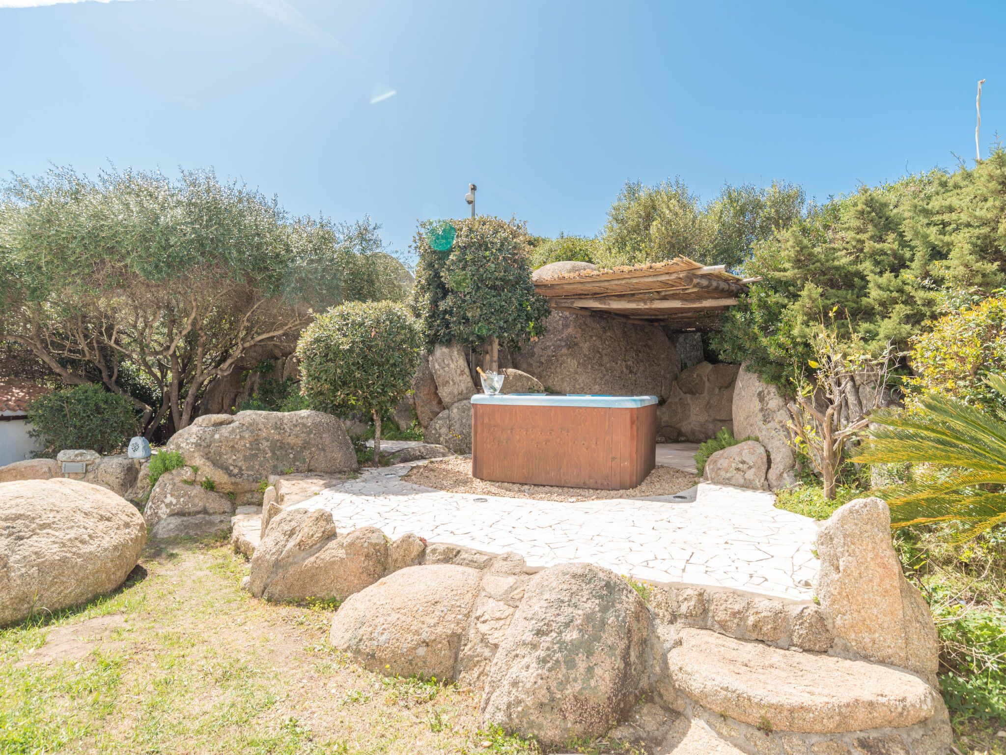Un bain à remous entouré de gros rochers et de verdure, avec un sentier de pierres menant à cet endroit.