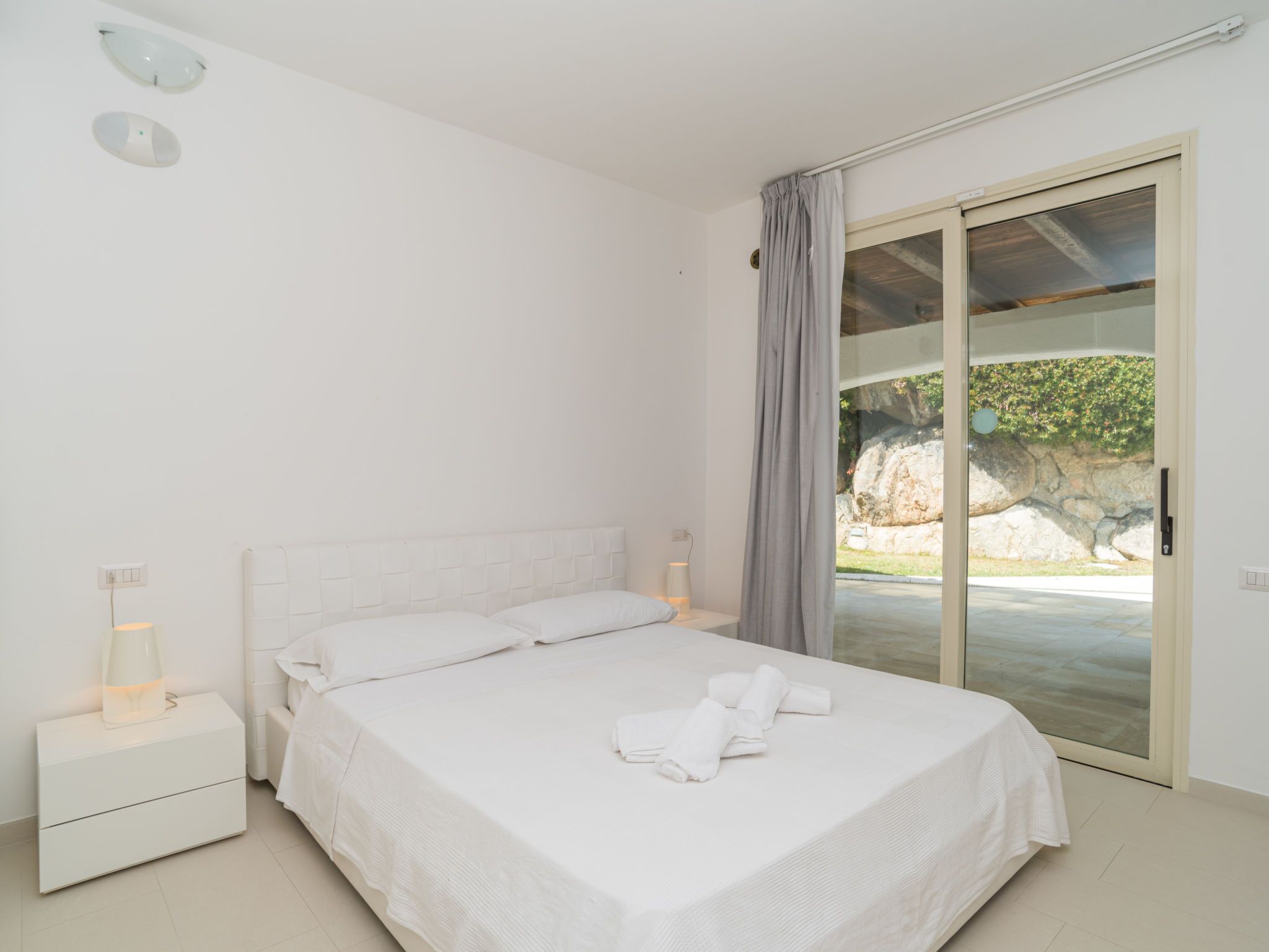 A bedroom with a bed, a nightstand, a lamp, and a view of a natural landscape through a sliding glass door.