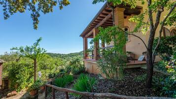 Una villa rustica con un vialetto in pietra che conduce a un pontile in legno, circondata da lussureggiante vegetazione e colline sullo sfondo.