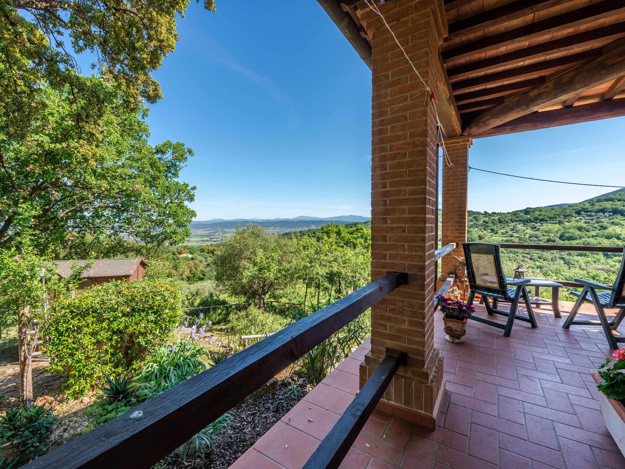 Un balcon agrémenté d’un pilier en briques, d’une balustrade en bois et offrant une vue sur un paysage luxuriant, avec des arbres et des collines à l’horizon.