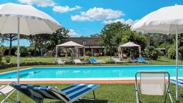 A swimming pool with lounge chairs and umbrellas, surrounded by greenery and a stone building in the background. - Le Cascatelle by Interhome (Massa Marittima)