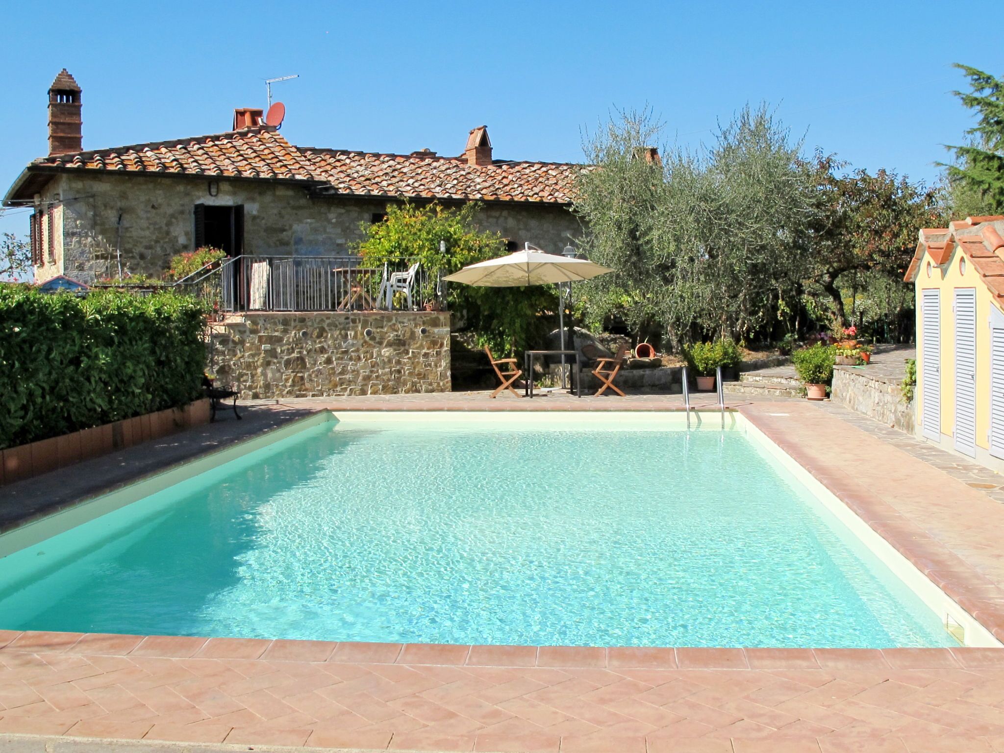 A rectangular swimming pool with a stone wall and tiled deck. There are two lounge chairs and a table under a canopy. A stone building with a tiled roof and chimney is visible in the background.
