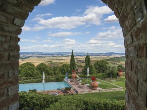 Une vue à travers une arche en pierre donnant sur un espace piscine, avec des chaises longues et des plantes en pot, et offrant un panorama sur un paysage pittoresque composé de collines ondulantes et d’un ciel dégagé.