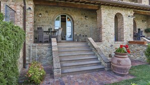 A stone building with a patio, outdoor seating, and a large planter with red flowers. - Appartamento Ginepro - Borgo in Chianni by Interhome (Chianni)