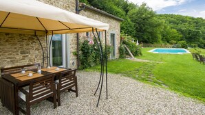 A stone building with a patio table and chairs under a canopy, a swimming pool in the background, and a grassy area with trees. - La Fonte by Interhome (Dicomano)