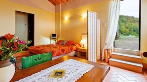 Una habitación con techo de madera, una cama con ropa de cama roja, una mesita de noche con una lámpara, una mesa con mantel de encaje y vistas al exterior con vegetación.