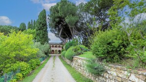 A gravel path leading to a building with arches, surrounded by lush greenery and trees. - Cellole - Torretta (CTC153) by Interhome (Castellina in Chianti)
