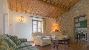 Uma sala de estar aconchegante com um sofá de linho verde xadrez, uma poltrona branca e uma mesa de centro de madeira. Uma parede de pedra e uma cozinha com armários verdes são visíveis ao fundo.