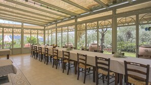 Ein überdachter Essbereich mit langem Tisch, gedeckt für eine Mahlzeit, umgeben von Stühlen und großen Fenstern mit Blick ins Freie.
