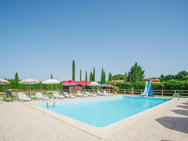 A swimming pool with lounge chairs and umbrellas, surrounded by greenery and a clear sky.