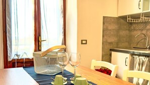 A dining area with a wooden table set for two, a kitchen with a sink and cabinets, and a window with curtains. - Apartment Ambra by Interhome (Pastina)