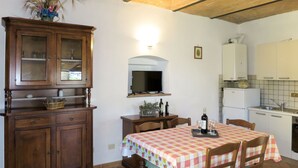 Ein Essbereich mit kariertem Tischlein, Holzstühlen, einer Schrank mit Glastüren, einem Fernseher über einem Holzregal und einer Küchenzeile mit weißen Schränken und einem Spülbecken.
