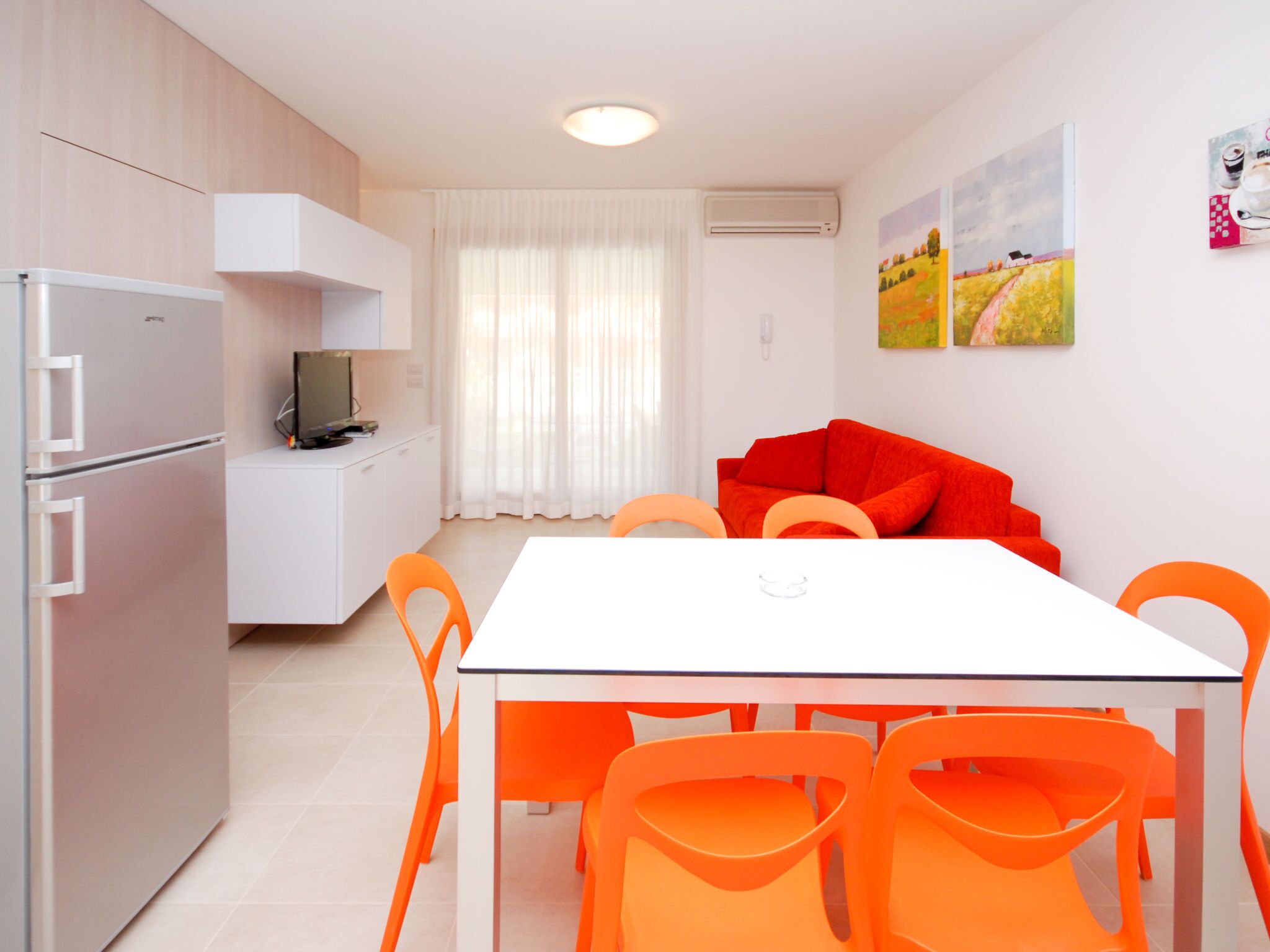 A modern kitchen with a refrigerator, stove, and microwave. There is a dining table with orange chairs and a red sofa. A painting is on the wall, and an air conditioner is mounted above the window.
