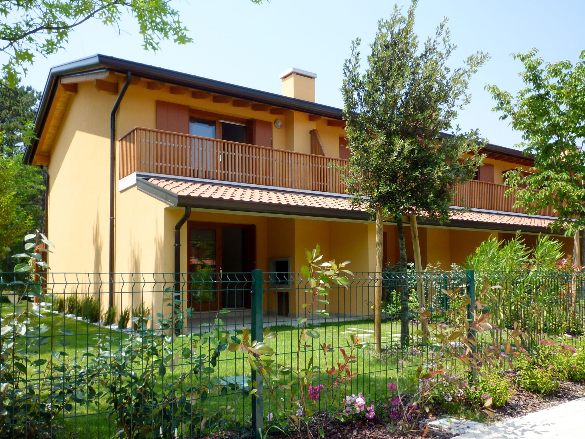 Ein zweigeschossiges Haus mit Balkon, umgeben von einem grünen Zaun und verschiedenen Pflanzen.