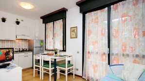 Quarto compacto com cama, mesa de jantar com cadeiras, pequeno espaço de cozinha e janela com cortinas florais.