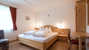 Una cama de madera con ropa de cama blanca, una mesita de noche pequeña, una silla y un armario de madera en una habitación con pisos de madera y paredes blancas.
