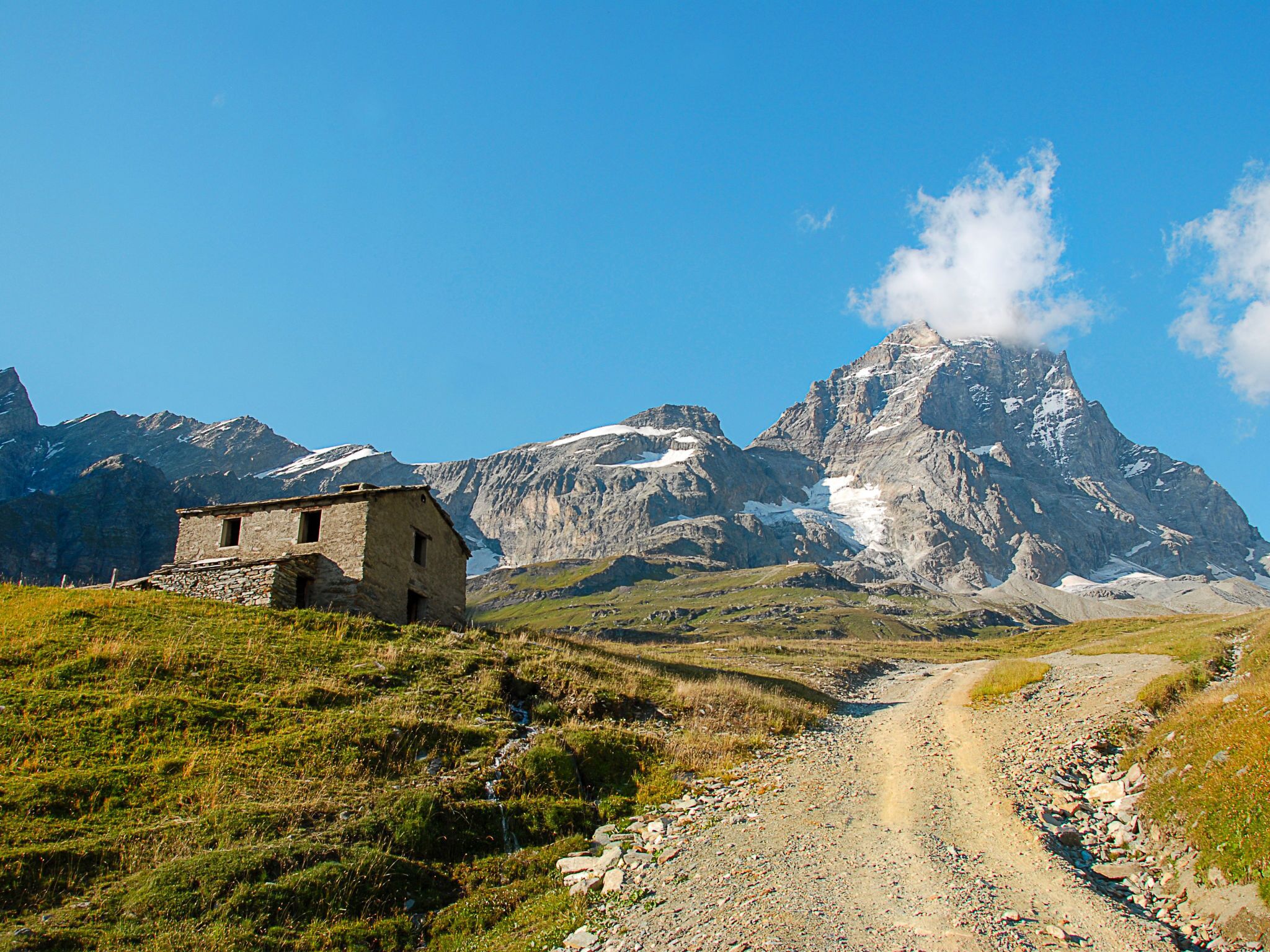 Un edificio de piedra rústico en una colina, con un sendero de tierra que lleva hacia una cordillera.