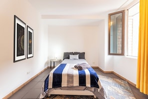 A bedroom with a bed, a nightstand, a window with curtains, and framed artwork on the wall.