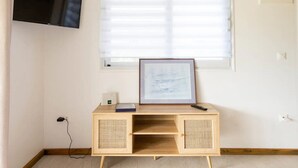Une pièce avec un téléviseur fixé au mur, une fenêtre avec des stores, un tableau encadré posé sur un support en bois et un thermostat au mur.