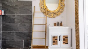 Une salle de bain aux murs recouverts de carreaux foncés, un meuble-lavabo blanc avec des portes en bois, un grand miroir doré à motifs et un escabeau en bambou.