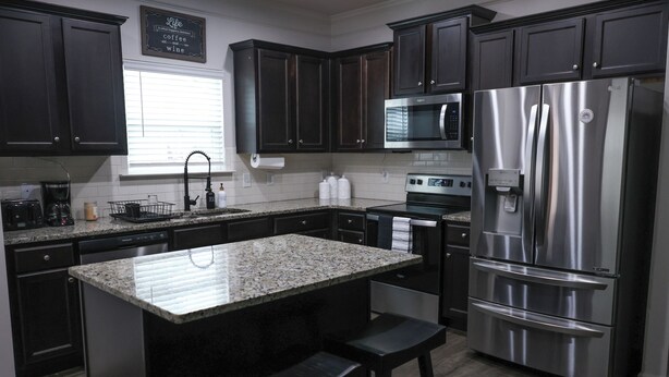 A modern kitchen with dark cabinets, stainless steel appliances, a granite island with bar stools, and a window with blinds. - Pet-friendly home minutes from Downtown/UGA Campus (Athens)
