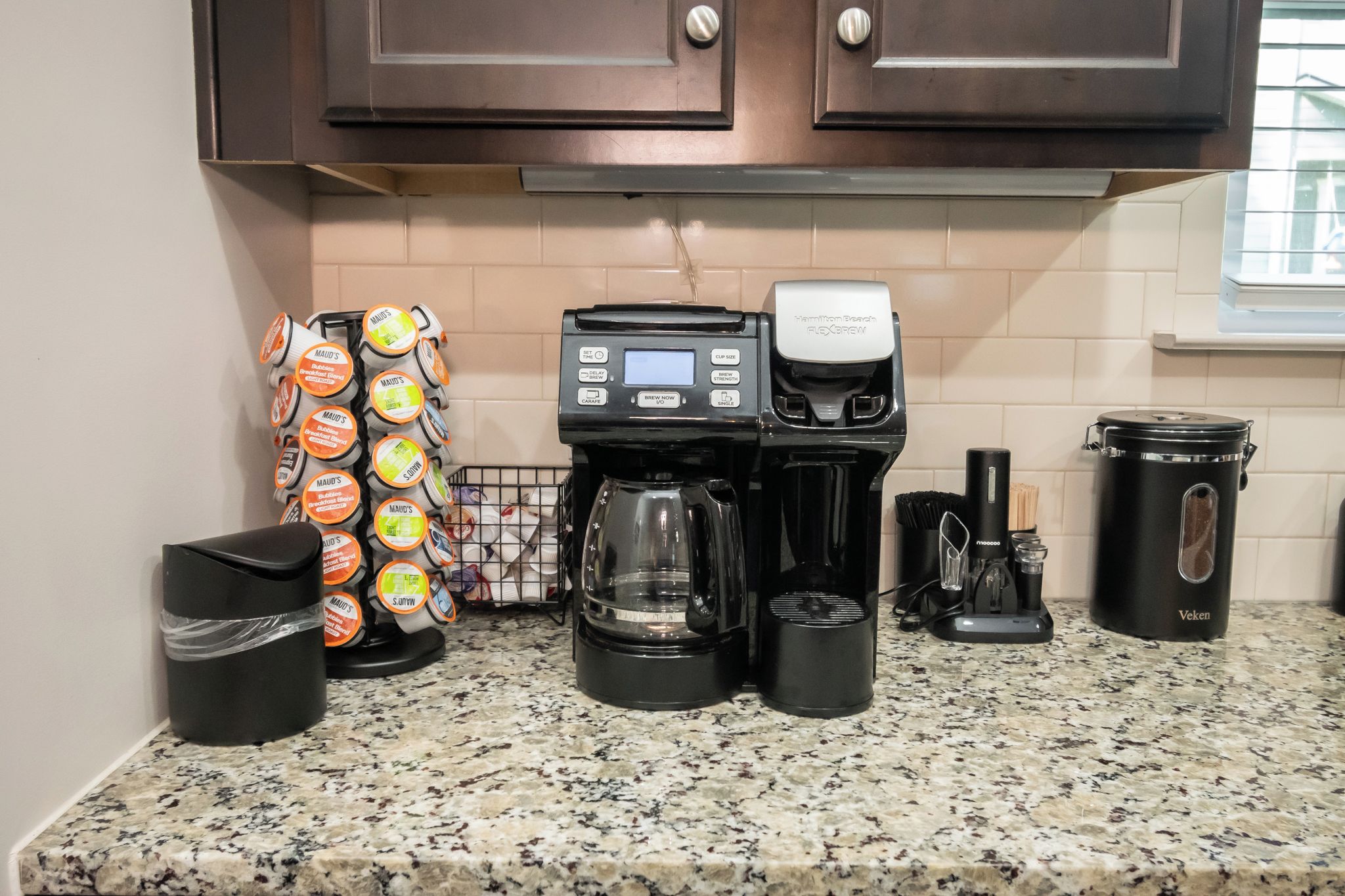 Un comptoir de cuisine avec une machine à café, une poubelle, un moulin à café et un récipient contenant des capsules de café.
