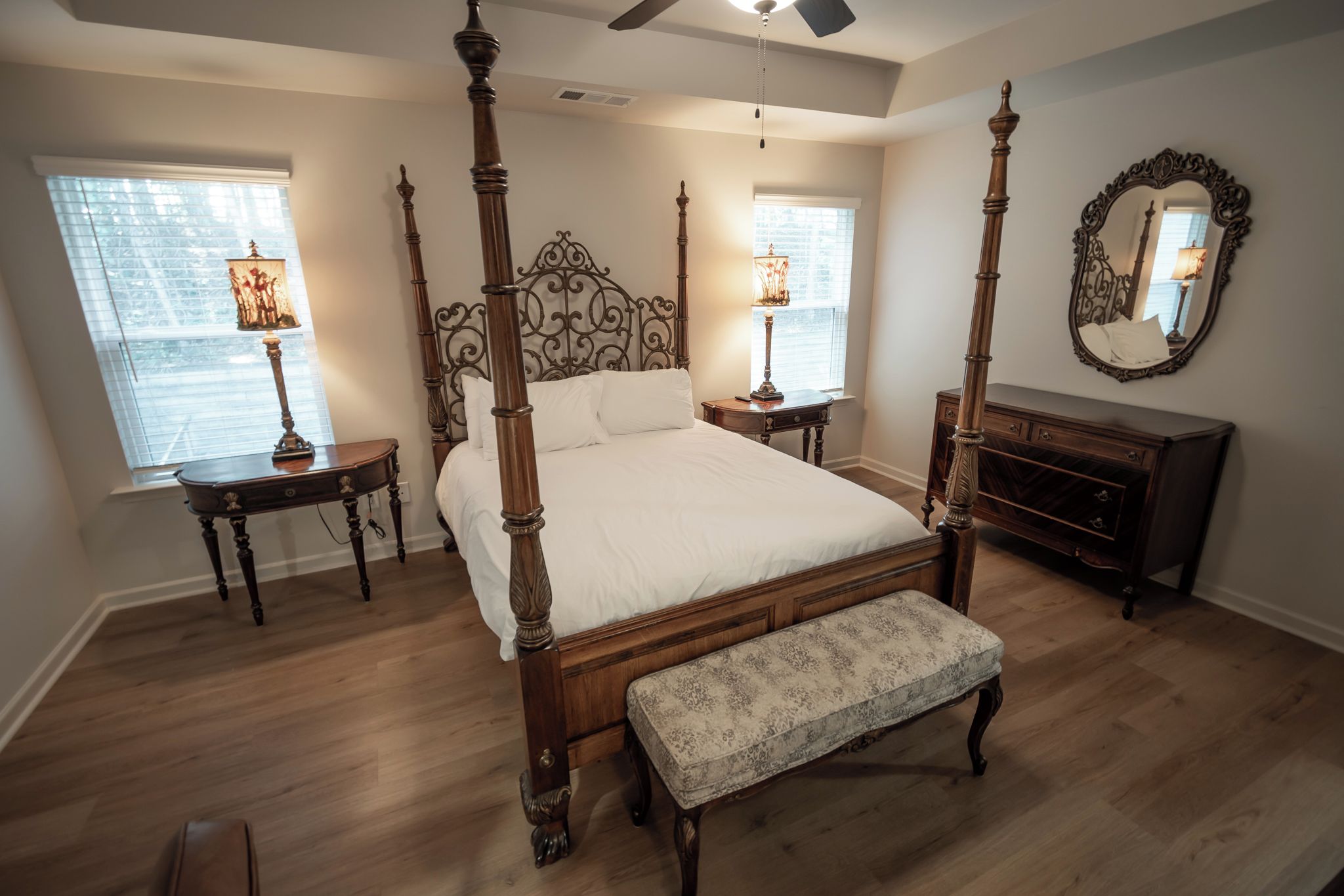 Un lit à baldaquin avec une tête de lit en bois, deux tables de chevet avec des lampes, un banc et une commode avec un miroir.