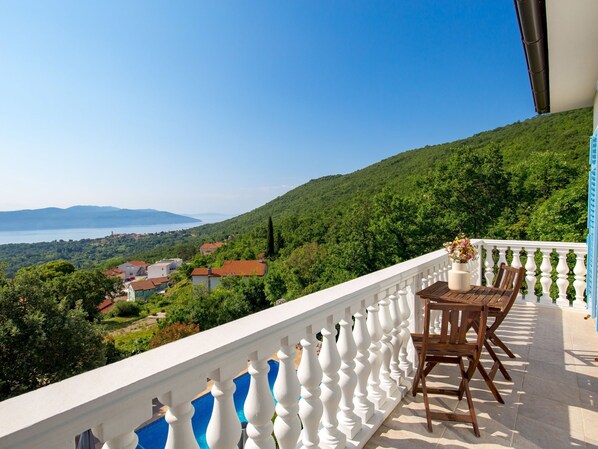 A balcony with a white balustrade, a wooden table with two chairs, and a view of a lake and hills.