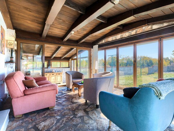 A spacious room with stone flooring, wooden beams, and large windows offering a view of the outdoors.