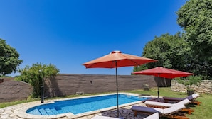 Una piscina con lettini e ombrelloni, circondata da piastrelle in pietra e vegetazione.