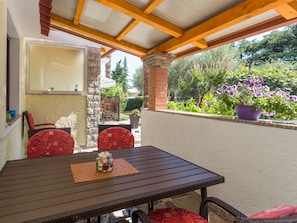 A covered patio with a wooden table and red chairs, a stone wall, and a view of a garden with flowers and greenery. - Apartment Nena by Interhome (Poreč)