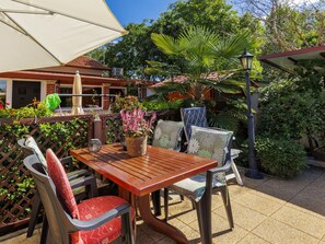 Un patio exterior con mesa y sillas de madera, rodeado de plantas y un paraguas blanco.