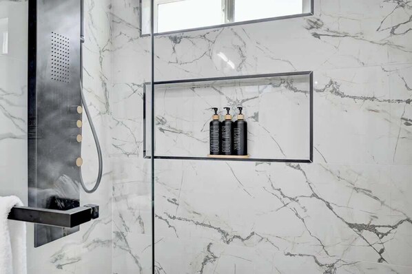 Un baño con pared de mármol, ducha con panel de control negro y tres botellas negras en un estante.