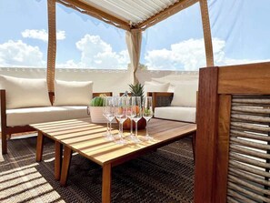 Una mesa de madera con copas de vino, un sofá y una planta en maceta en un área de estar al aire libre techada.