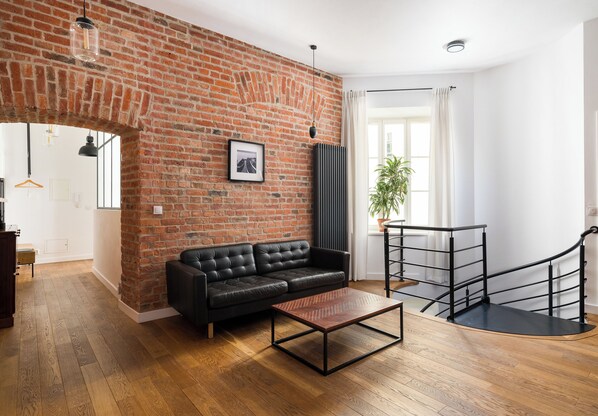 A living room with a brick wall, a black leather sofa, a wooden coffee table, and a staircase with a black railing.