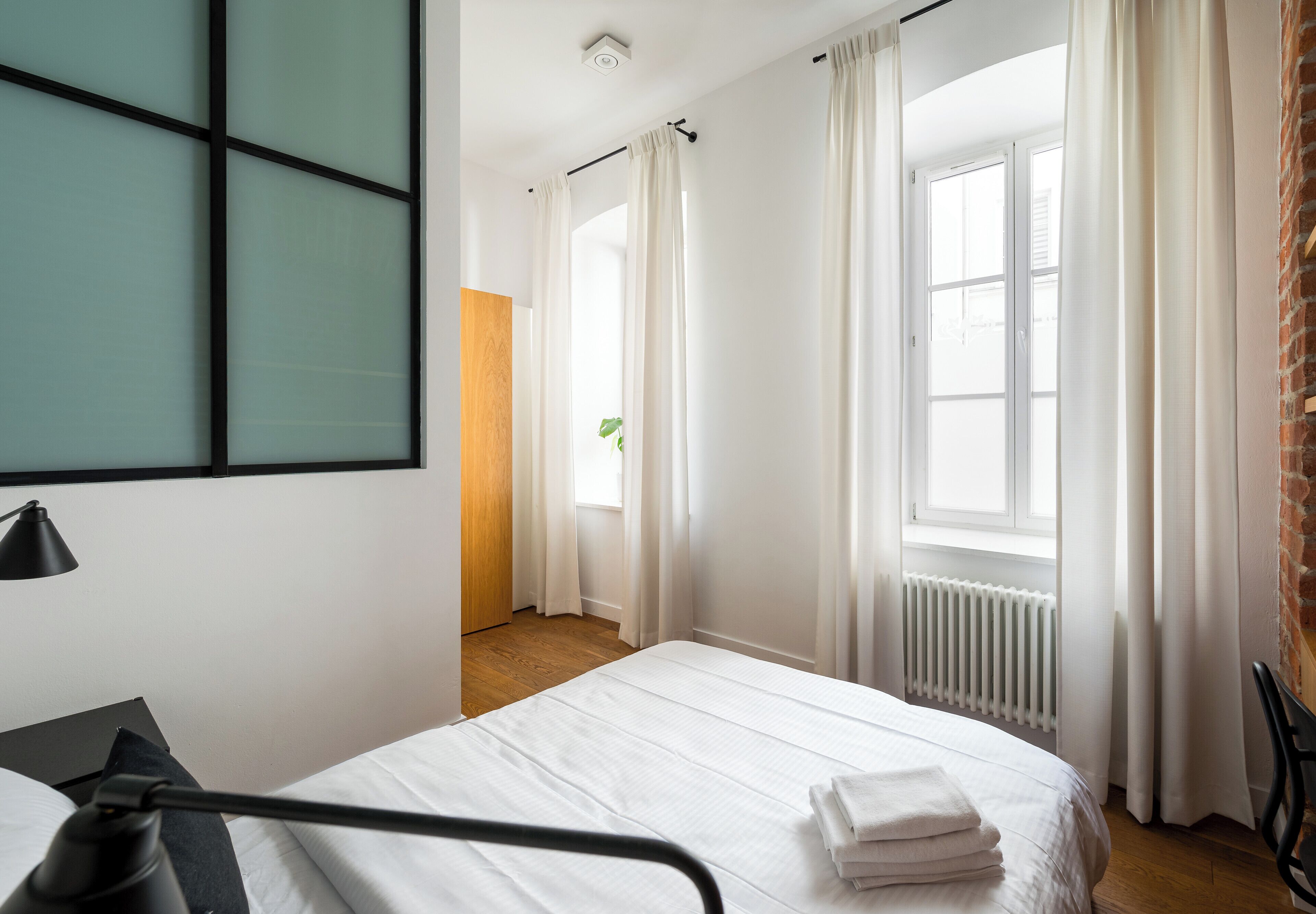 A hotel room with a bed, a desk, a chair, a radiator, and a window with curtains.