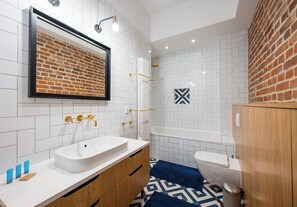 A modern bathroom with a white sink, wooden vanity, a large mirror, a shower with a gold showerhead, and a white toilet.