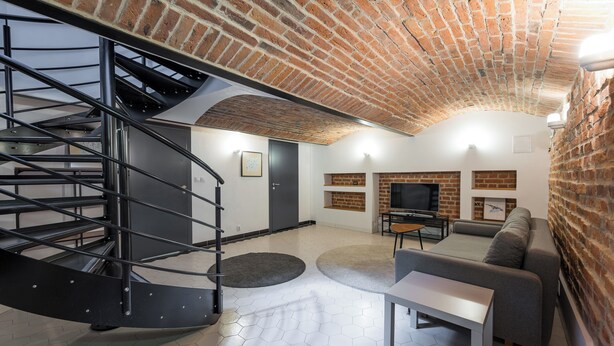A modern living room with a brick ceiling, a spiral staircase, a grey sofa, a TV, and a fireplace. - Park Praski Two-Level Apartment (Warszawa)