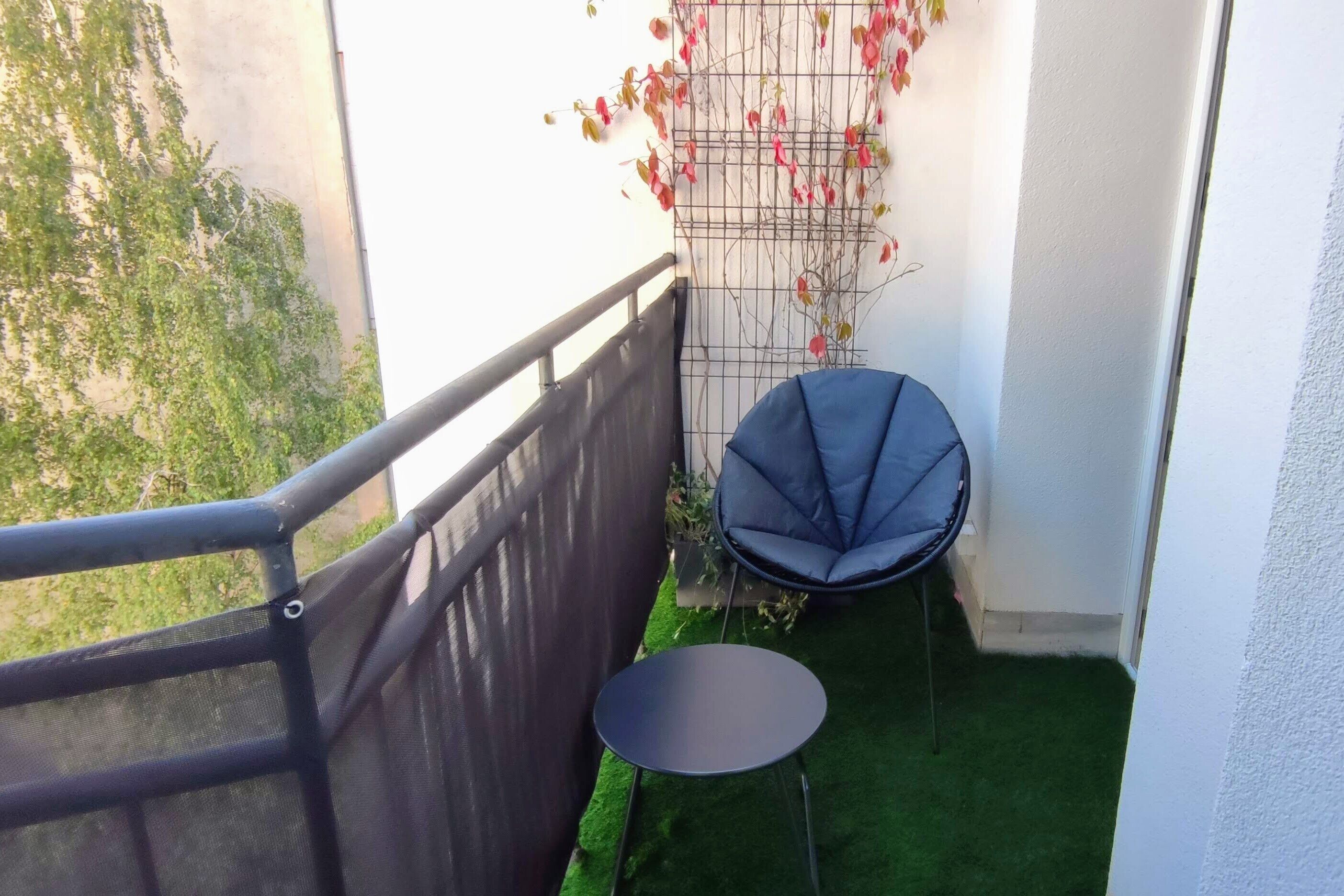 Un pequeño balcón con una silla negra y una mesa redonda, una pared blanca y un suelo de césped artificial verde.