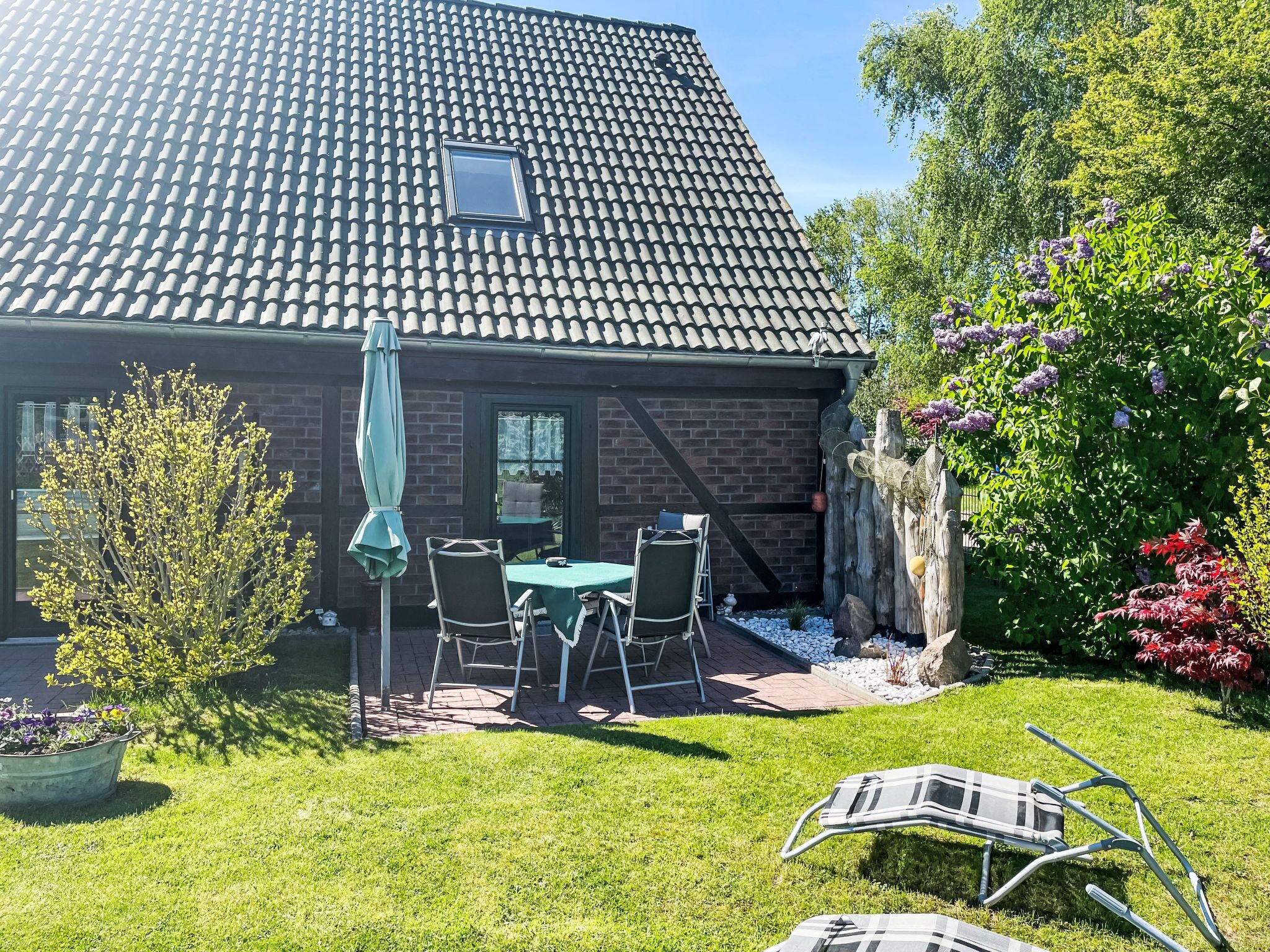 A house with a tiled roof, a brick exterior, a small patio with a table and chairs, and a garden with flowers and a tree.