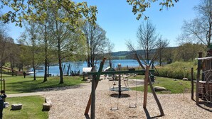 A playground with swings and a seesaw, surrounded by trees and a lake. - Alpenblick by Interhome (Illmensee)