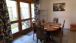Comedor con mesa de madera dispuesta para seis personas, rodeada de sillas. Hay un ventanal con cortinas, un cuadro en la pared y un estante pequeño.