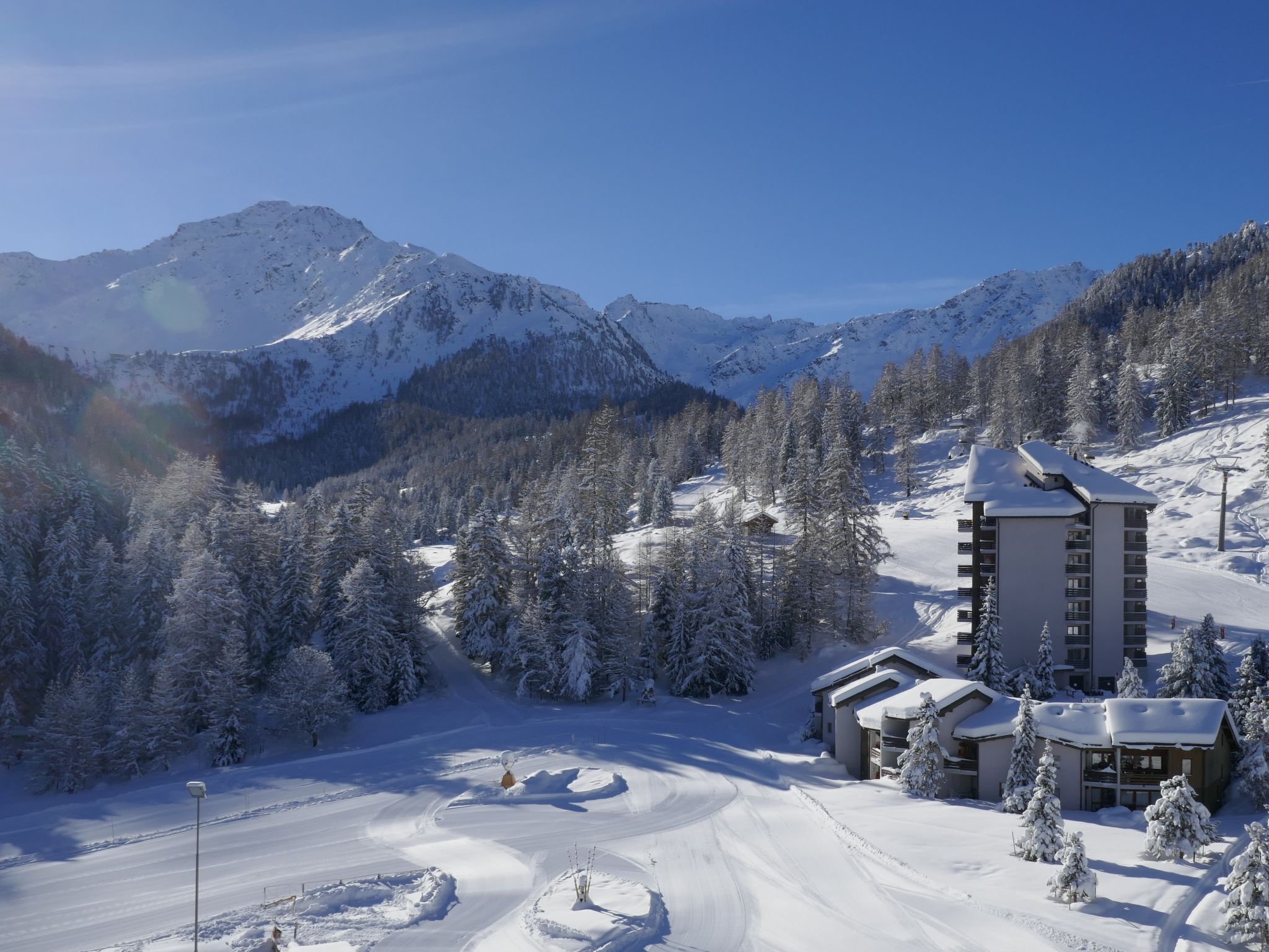 A snow-covered mountain resort with a ski slope, a tall building with multiple floors, and a forested area.